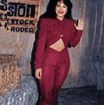 Selena Quintanilla posando con un conjunto rojo de dos piezas, sonriendo y saludando, con un fondo rústico que refleja su estilo vibrante y su energía