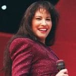 Selena Quintanilla sonriendo mientras sostiene un micrófono en el escenario, vistiendo un conjunto morado brillante, transmitiendo su energía y carisma durante una actuación en vivo.